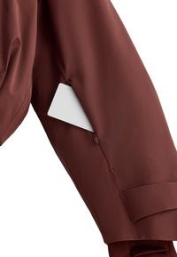 Barna vízálló dzseki anyag zipzáros zsebbel az ujjakon, amely félig felfedi a benne lévő fehér kártyát. Simább textúra, letisztult dizájn.