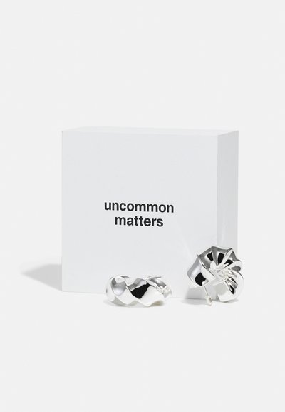 Cercei din metal argintiu cu un design răsucit și floral, așezați lângă o cutie albă simplă cu eticheta "uncommon matters" scrisă cu text negru.