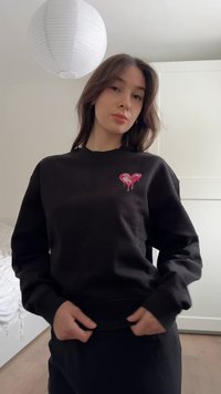 Νεαρή γυναίκα μέσα σε εσωτερικό χώρο φοράει μαύρο φούτερ με ροζ σχεδιασμό καρδιάς που στάζει, προσαρμόζοντας το τελείωμα, με λευκό χαρτί λάμπα πάνω από το κεφάλι της.