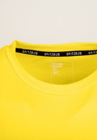 Gelbes Baumwoll-T-Shirt mit Rundhalsausschnitt, einem schwarzen Etikett mit der Aufschrift "SPITZBUB" und Pflegehinweisen auf der Innenseite.