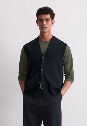 Marineblauwe gebreide vest met een v-hals, knopenfront en ribgebreide randen, gedragen over een olijfgroen langemouwen shirt, gecombineerd met donkere broek.
