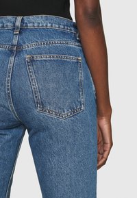 Jeans de mezclilla azul con cintura alta, que cuentan con dos bolsillos traseros y costuras en contraste. La tela tiene una textura y un aspecto ligeramente desgastado.
