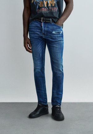 Uomo con jeans blu slavati a taglio slim con una toppa consumata, cintura nera con borchie e sneakers nere, in piedi con una mano in tasca.