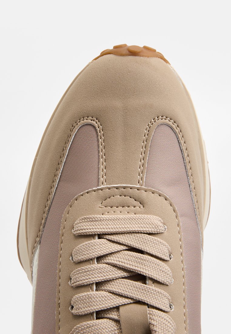 Zapatilla beige con un upper de ante suave, acentos de malla y una suela de goma texturizada. Presenta un diseño de cordones planos y detalles cosidos.