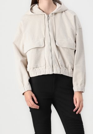 Kvinde iført en lysebeige corduroy hættejakke med lynlås, store forlommer og sorte bukser med ribbet knædetaljer.