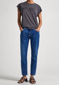 Šedé tričko s krátkým rukávem se nápisem "PEPE JEANS" tištěným černě, spárované s modrými rovnými džíny a černými sandály na podpatku se sponami.