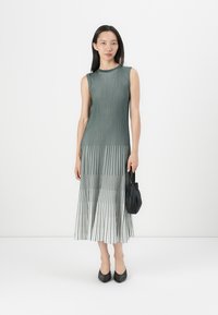 Pleated dress in green and white, sleeveless design with vertical stripes. Paired with a small black bag and black shoes. Simple silhouette.
