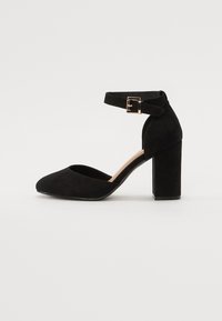 Svart mocka ankelremssko med spetstopp och en tjock blockklack. Har en metallspänne på remmen och en slät yta.