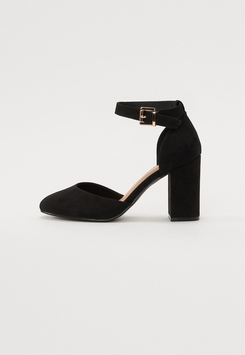 Svart mocka ankelremssko med spetstopp och en tjock blockklack. Har en metallspänne på remmen och en slät yta.