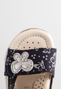 Gros plan d'une sandale beige avec une bride noire décorée d'une broderie papillon argentée et de petits points argentés.