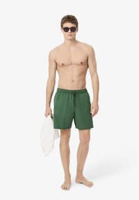 Shorts de bain verts en tissu léger, dotés d'une taille élastique avec cordon de serrage. Le mannequin tient une serviette blanche.