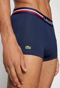 Marineblaue Boxershorts mit einem gerippten Bund, der rote und weiße Streifen aufweist. Lacoste-Logo in Grün auf der unteren linken Seite. Weicher, elastischer Stoff.