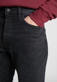 Mustat farkut, joissa on vartalonmyötäinen leikkaus, edessä tasku, hillitty ompelu ja nappikiinnitys. Yhdistetty viininpunaiseen collegepaitaan.