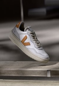 White sneaker featuring a textured mesh upper, tan leather accents, and a thick cream rubber sole. Lace-up design with logo detail on the tongue.