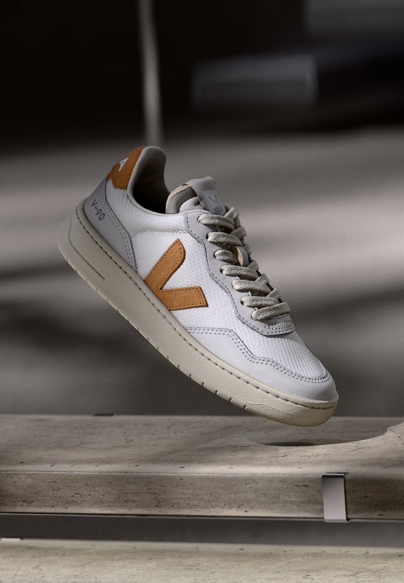 White sneaker featuring a textured mesh upper, tan leather accents, and a thick cream rubber sole. Lace-up design with logo detail on the tongue.