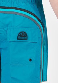 Pantaloni da bagno turchesi con tasca laterale, caratterizzati da strisce di accento nere e azzurro chiaro e un patch logo SUNDEK nero. Texture liscia.