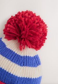 Strikket hue med rød kvast, med blå og hvide striber. Blød garntekstur, rund form og kontrasterende farveaccenter.