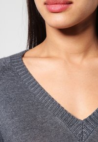 Grauer, gestrickter Pullover mit V-Ausschnitt, der über ribbelnde Details am Kragen und eine weiche Textur verfügt. Nahaufnahme des Kragenbereichs.