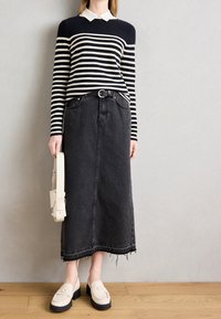 Kobieta w czarno-białym swetrze w paski, czarną spódnicą jeansową midi z postrzępionym dołem, beżową torbą nerka i beżowymi mokasynami z czarnymi podeszwami.