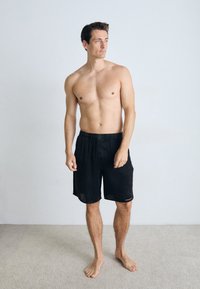 Black elastic waistband shorts, knee-length, lightweight fabric, featuring a small accent detail. Barefoot model with a neutral background.