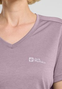 T-shirt lavanda a scollo a V realizzato in tessuto morbido, con un piccolo logo bianco Jack Wolfskin sulla parte sinistra del petto.