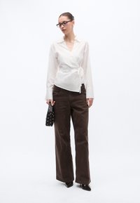 Λευκό μπλουζάκι τύπου wrap με λεπτομέρεια δεσίματος, συνδυασμένο με σκούρο καφέ παντελόνι ευρείας πατούρας. Αξεσουάρ περιλαμβάνουν μια μαύρη τσάντα με υφή και τρουκς και παπούτσια με τακούνι.