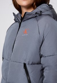 Szara kurtka puchowa z kapturem, z przednim zamkiem błyskawicznym, kieszeniami bocznymi oraz haftowanym logo w kolorze pomarańczowym na klatce piersiowej. Gładka tekstura.