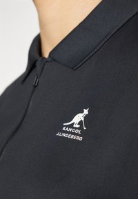 Juodos polo marškiniai su nėriniuotu apykakle ir baltu prekės ženklo logotipu, vaizduojančiu kengūrą, esantį kairėje krūtinės pusėje.