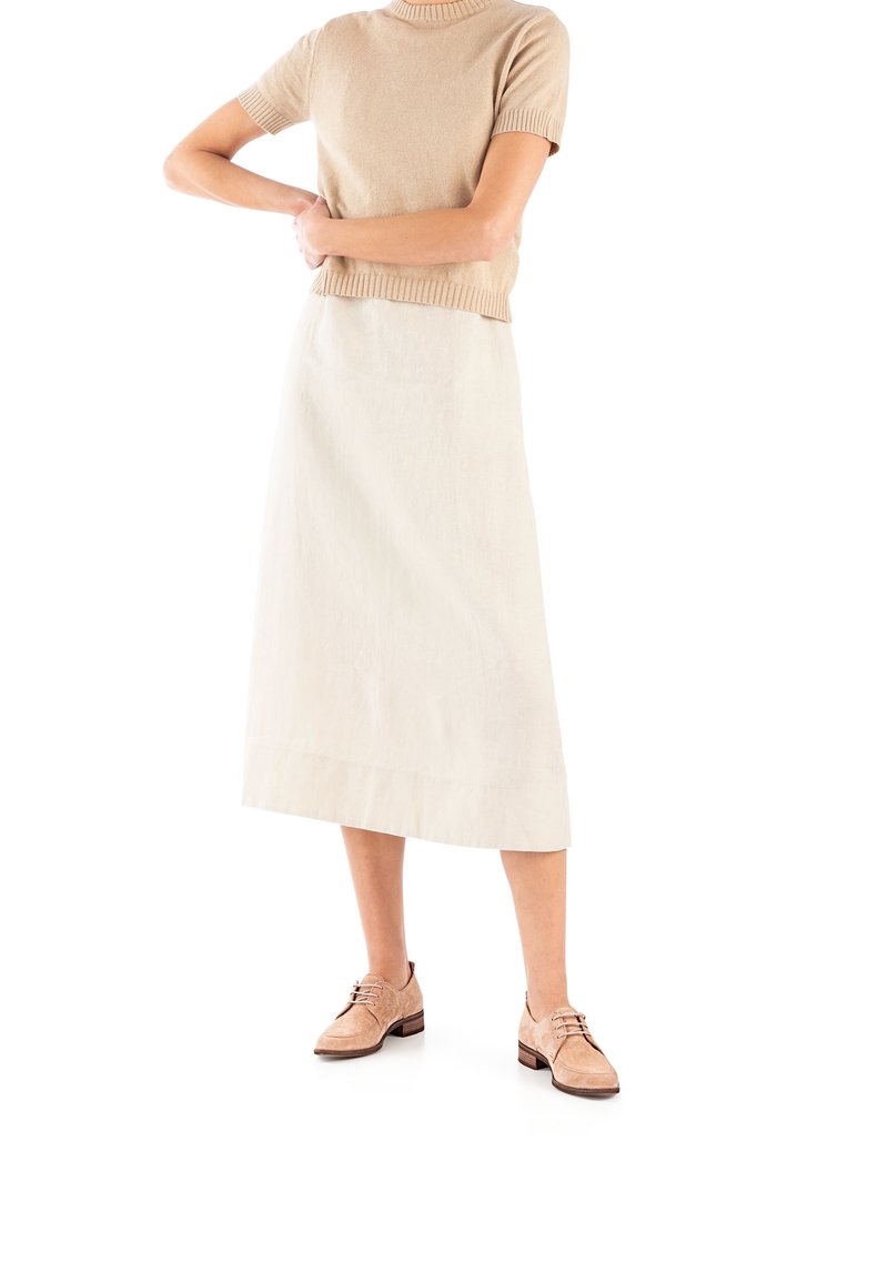 Femme portant un pull beige à manches courtes, une jupe midi blanche et des chaussures à lacets marron clair, debout, les bras croisés à la taille.