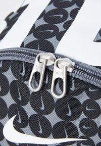 Σακίδιο φτιαγμένο από παλιό μαύρο και γκρίζο υφασμάτινο σχέδιο με ασημένιους φερμουάρ, που διαθέτει το λευκό λογότυπο της Nike και στρογγυλούς λογότυπους ως αξεσουάρ.