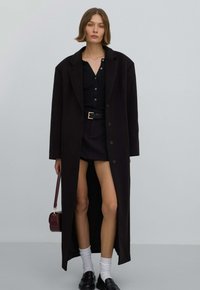 Lange zwarte jas, nauwsluitende zwarte top en korte zwarte rok. Zwarte schoenen, witte sokken en een klein bordeauxrood handtasje maken de look compleet.