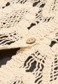 Kreemika heegeldatud kangas, mis sisaldab lillemustreid, keerukaid tekstuure ja naturaalset puitnööpi tumedal taustal.