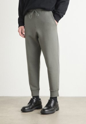 Pantalons de jogging vert olive avec une taille élastique et un cordon de serrage, présentant une coupe slim et des poignets côtelés. Associés à des chaussures noires à lacets.
