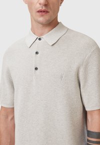 Lichtgrijs gebreid polo shirt met een gestructureerd patroon, korte mouwen, een klassieke kraag en drie zwarte knopen aan de voorkant. Subtiele merkdetails.
