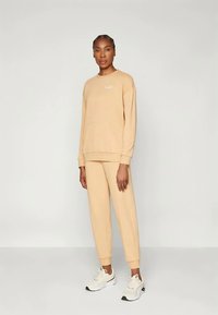 Beżowa bluza i dopasowane spodnie dresowe wykonane z miękkiej tkaniny. Bluza ma okrągły dekolt i haftowane logo; spodnie dresowe posiadają elastyczne mankiety.