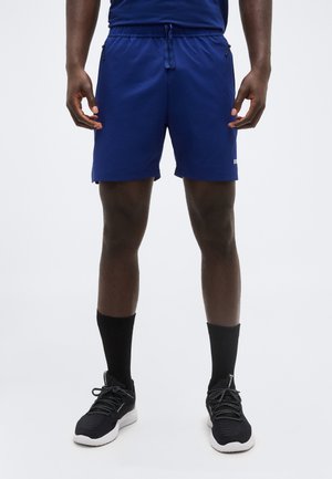 Man draagt blauwe sportkorte broek, zwarte sokken en zwarte hardloopschoenen en staat met ontspannen handen langs zijn lichaam.