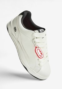 Valkoiset tennarit, joissa on musta sisävuori, valkoiset nauhat ja punavalkoinen koiralogo-Tag, joka roikkuu nauhoista valkoisella taustalla.