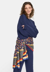 Kobieta z kręconymi włosami, ubrana w granatowy strój i kolorowy, wzorzysty szal przewiązany w talii, stoi z rękami skrzyżowanymi.