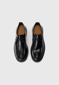 Zapatos de charol negros de piel con diseño slip-on, que cuentan con una puntera redondeada, superficie lisa y un sutil detalle de costura. La suela de goma plana completa el diseño.