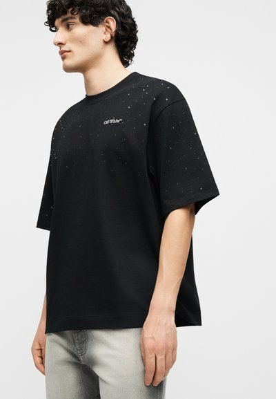 Νεαρός άνδρας με σγουρά μαλλιά που φοράει μαύρο T-shirt Off-White με μικρές λευκές κηλίδες και ανοιχτό γκρι τζιν, κοιτάζοντας προς τα αριστερά.