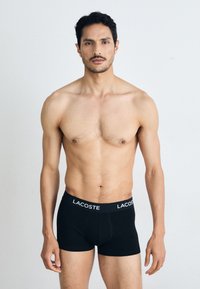 Black boxer briefs with a Lacoste logo on the waistband, fitted design, soft fabric, and classic cut, worn by a standing male model.