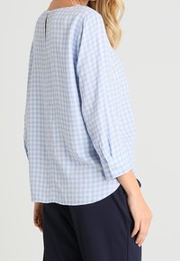 Hellblau-weiß kariertes Blusenshirt mit rundem Halsausschnitt, drei Viertelärmeln und einer kleinen Öffnung auf der Rückseite, aus einem weichen Stoff gefertigt.