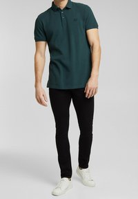 Groen poloshirt met korte mouwen, drieknoopsluiting en een klein logo. Gecombineerd met zwarte slim-fit jeans en witte sneakers.