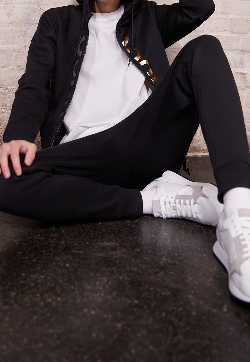 Felpa nera con zip e dettagli dorati, t-shirt bianca, pantaloni della tuta neri e sneakers grigie/bianche, seduto su un pavimento scuro e strutturato.