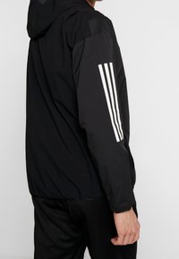 Svart sportjacka med huva, texturerat tyg och tre vertikala vita ränder på vänster ärm. Enfärgad svart design.