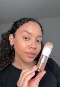 Maybelline foundation stick with a pink cap, clear body, and light brown product. Curly hair, silver hoop earring, wearing a black shirt.