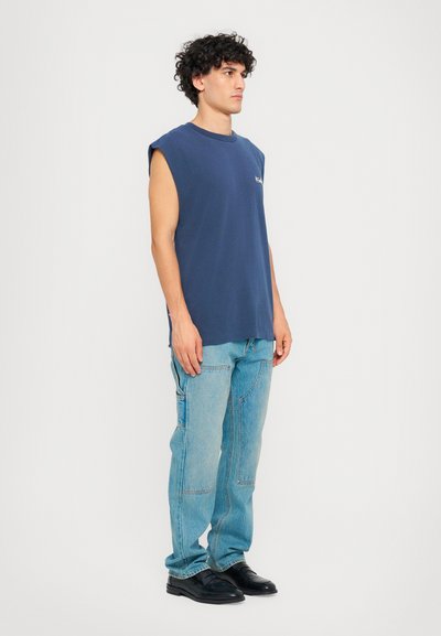 Tricou fără mâneci bleumarin din bumbac, cu o croială lejeră și guler rotund. Asortat cu blugi din denim deschis la culoare, cu cusături vizibile și buzunare laterale.