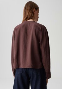 Woman with long brown hair wearing a loose, long-sleeve brown top and dark blue jeans, standing against a plain light background.
