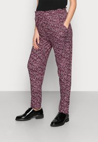 Woman wearing burgundy patterned tapered pants and black patent leather lace-up shoes, standing with one hand in pocket.