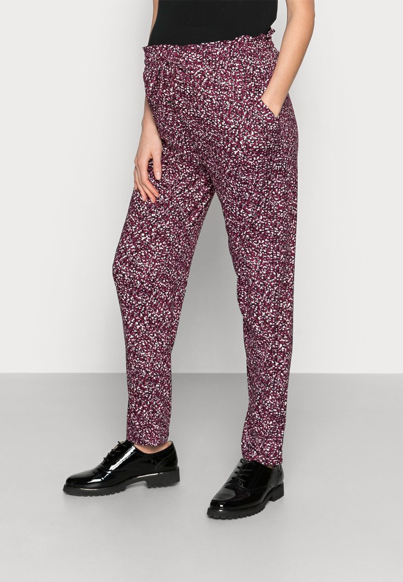 Woman wearing burgundy patterned tapered pants and black patent leather lace-up shoes, standing with one hand in pocket.